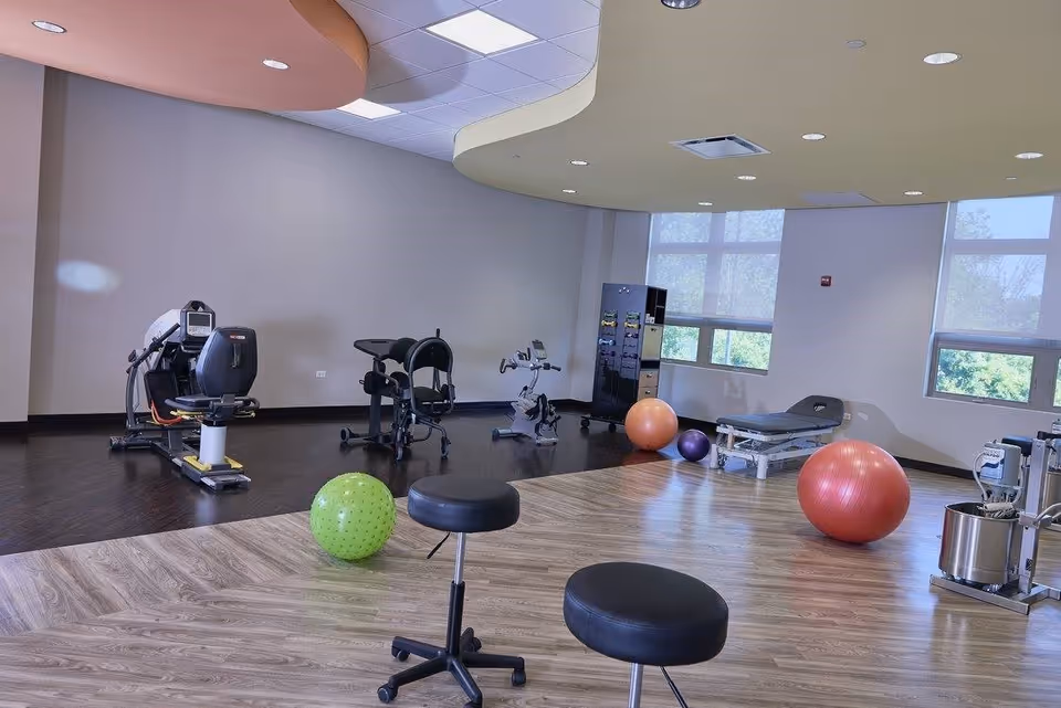 A spacious exercise and rehabilitation room with various fitness equipment including stationary bikes, exercise balls in green, orange, and purple, two black stools on wheels, a treatment table, and a storage cabinet. Large windows allow natural light to fill the room.