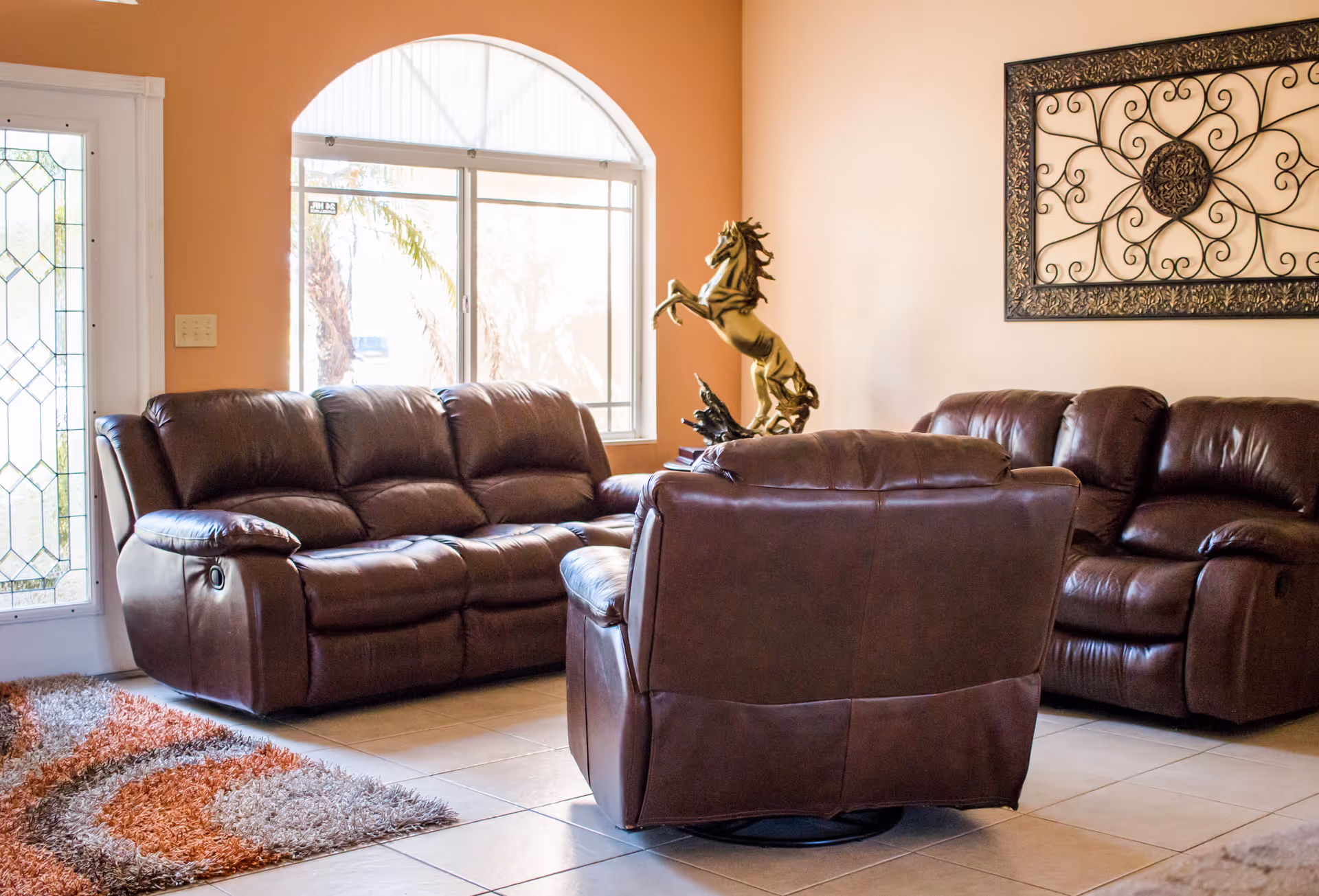 A living room with brown leather sofas and a matching armchair arranged around a central area. There is a decorative metal wall art piece on the beige wall and a golden horse statue on a table near the window. The room has a tiled floor and a door with decorative glass panels. A multicolored rug is partially visible on the floor.