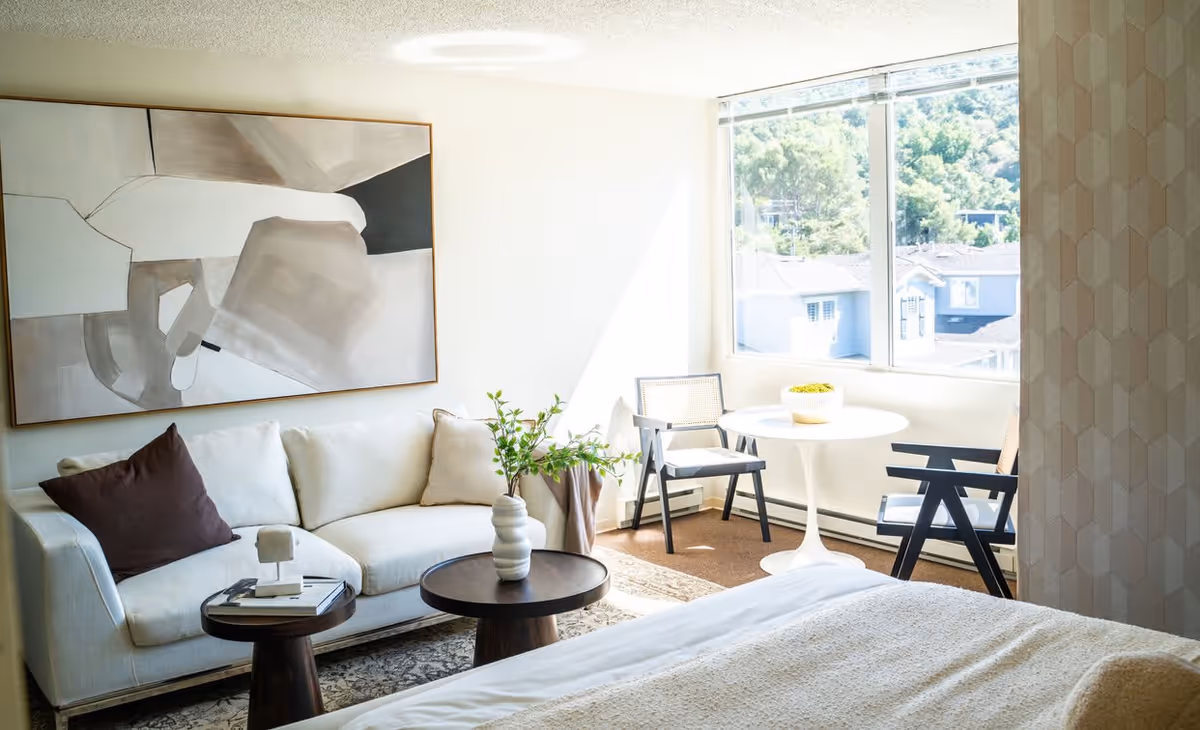 Sunlit studio-style living area with a sofa, coffee tables, a bed in the foreground and a small round dining table with chairs by a large window.