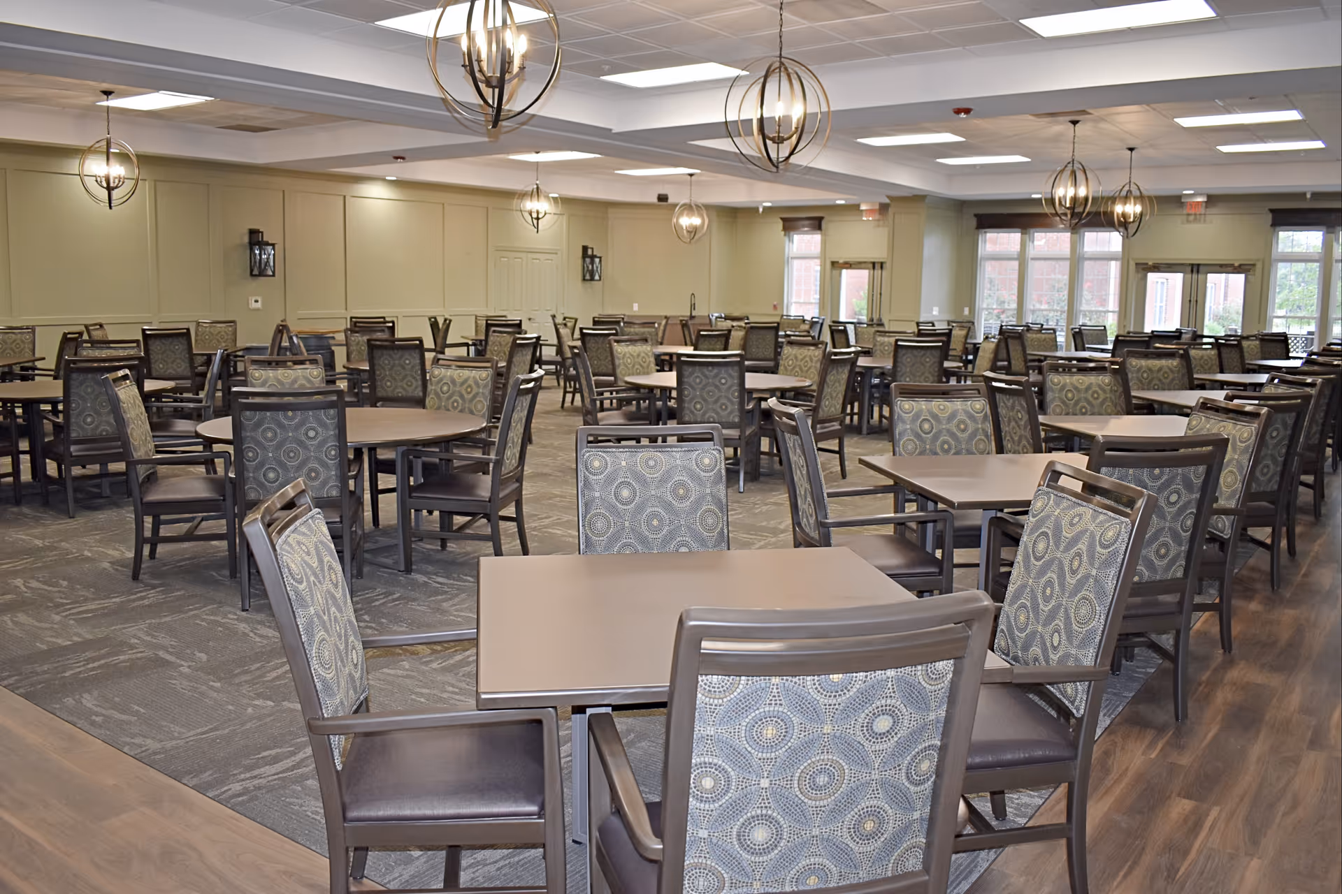A spacious dining room with multiple square and round tables surrounded by patterned cushioned chairs. The room features modern spherical pendant lights hanging from the ceiling, large windows allowing natural light, and a combination of carpet and wood flooring.