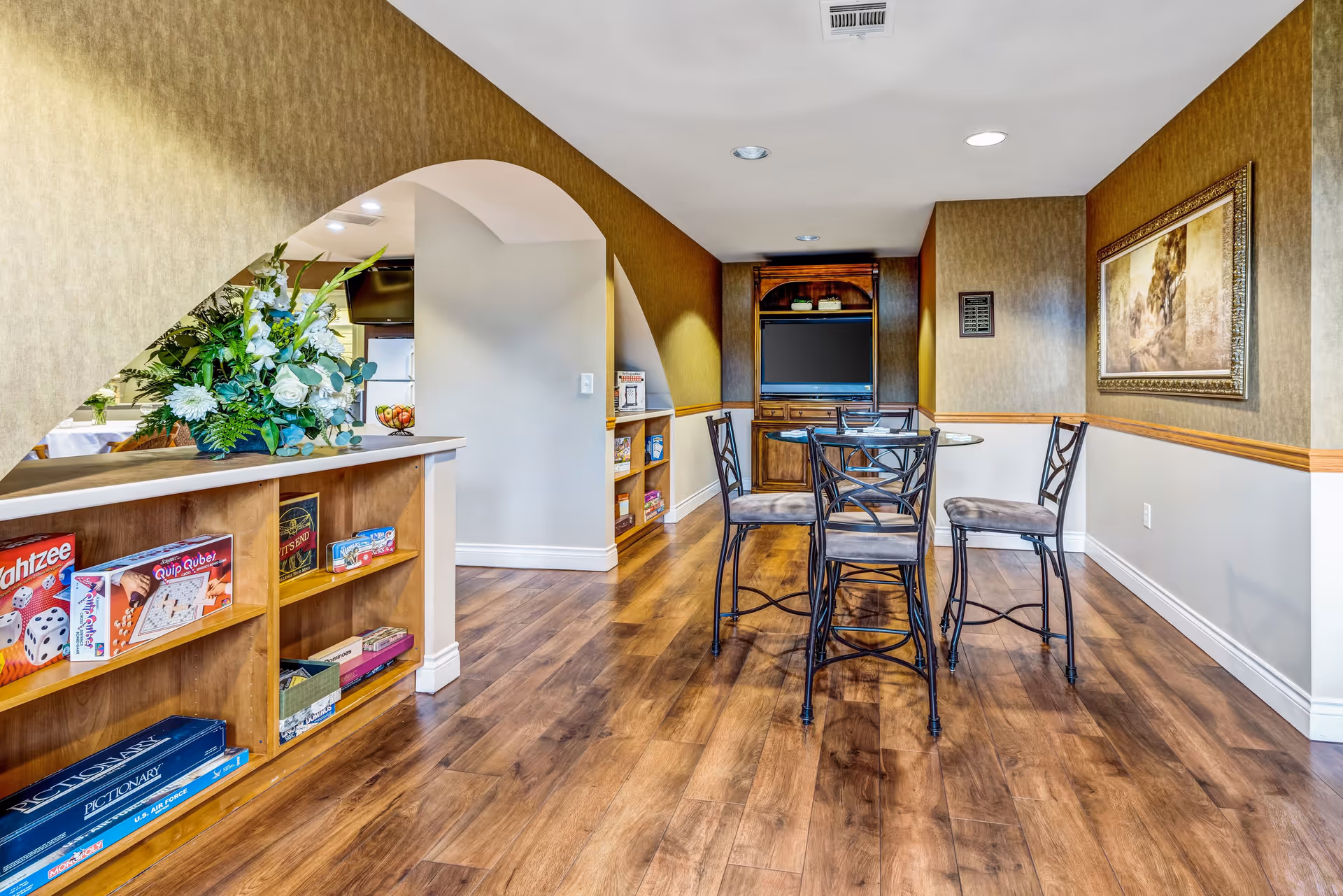A cozy interior common area with wooden flooring, a small round table with four chairs, built-in wooden shelves holding board games, a floral arrangement on a counter, and a wall-mounted TV in a wooden cabinet. The walls are decorated with a framed painting and have a two-tone color scheme with a chair rail.
