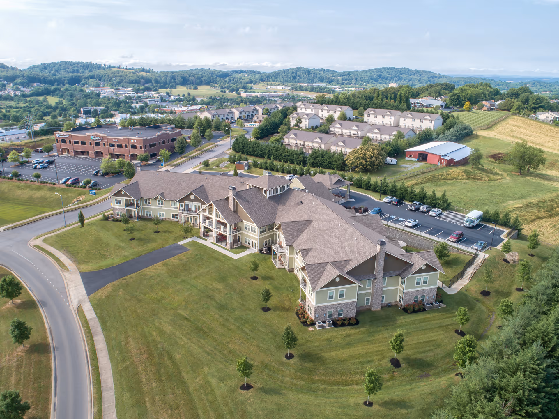 Aerial view of Dominion Senior Living facility showing a large, multi-wing building surrounded by well-maintained lawns, small trees, and parking lots. The building has a beige exterior with stone accents and multiple balconies. The surrounding area includes other buildings, roads, and green hills in the background under a partly cloudy sky.