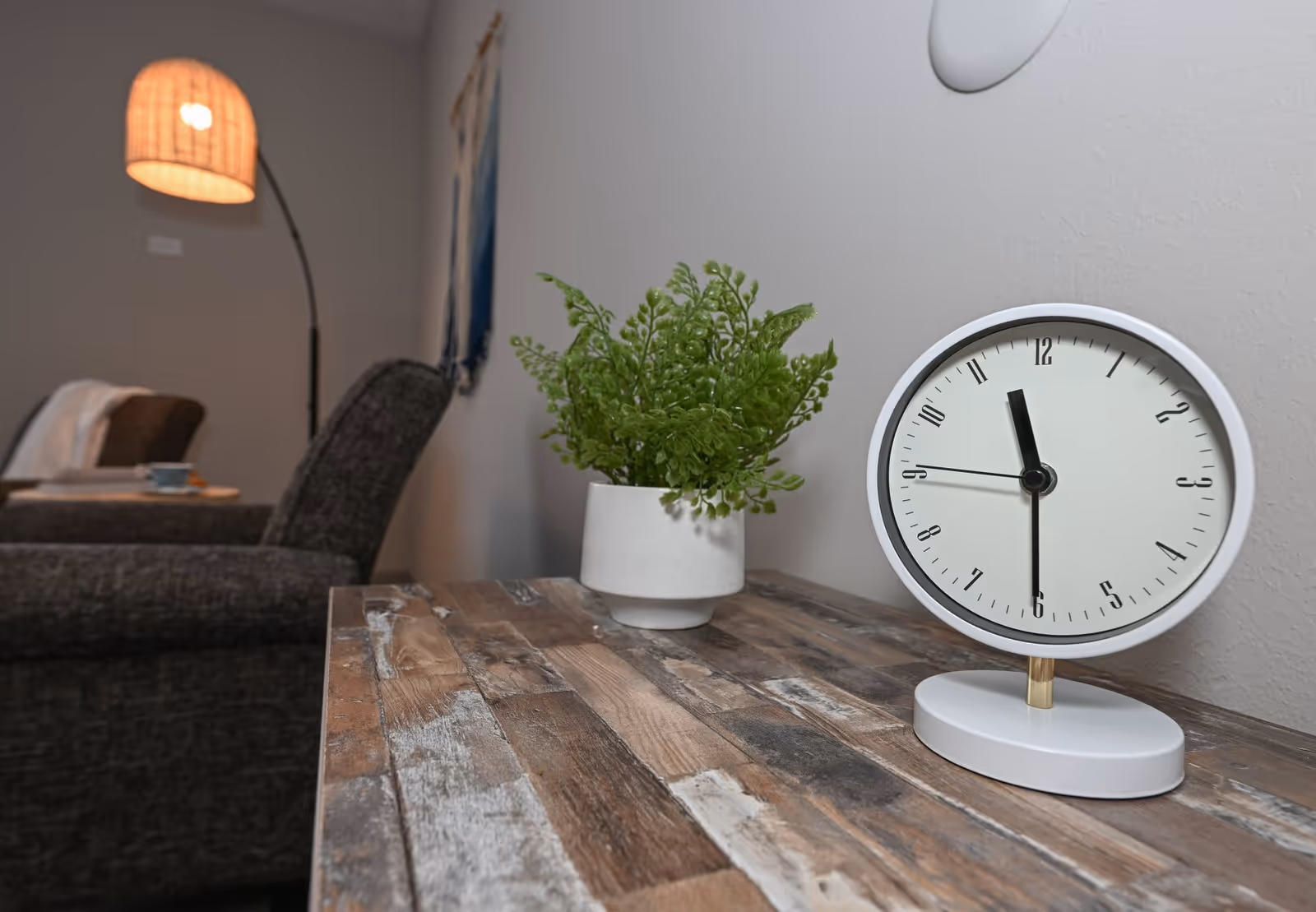 A close-up view of a rustic wooden table with a white round clock showing 11:30 and a small potted green plant. In the background, there is a gray upholstered chair, a floor lamp with a woven lampshade, and a wall hanging.