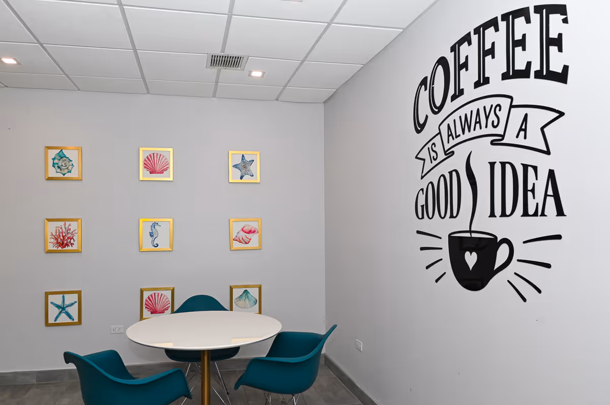 A small seating area with a round white table and three teal chairs. The wall on the right has a large black decal that reads 'Coffee is always a good idea' with a steaming coffee cup illustration. The wall on the left has nine framed pictures of sea-themed items like shells, starfish, and coral arranged in a grid.