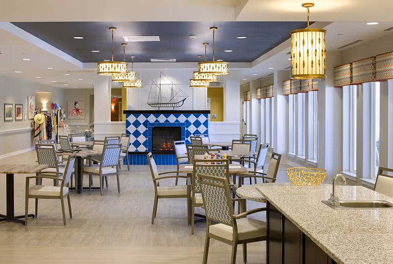 A spacious dining area with multiple tables and chairs arranged neatly. The room features a decorative fireplace with blue and white tiles and a model ship on the mantel. Large windows with patterned valances allow natural light to fill the space. Several pendant lights hang from the ceiling, and a kitchen island with a sink and a decorative bowl is visible in the foreground.