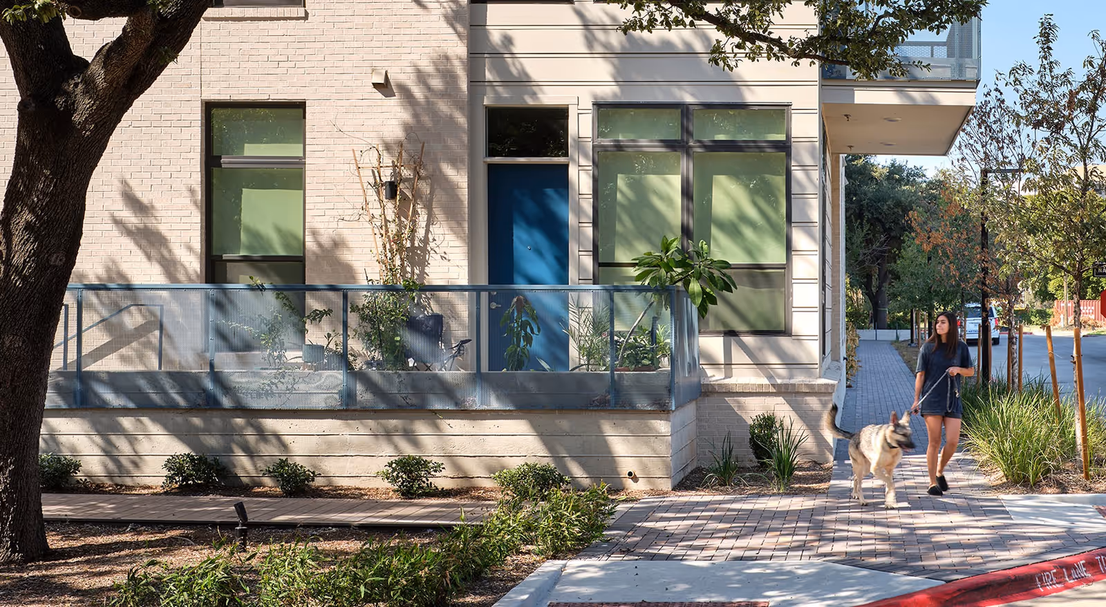 A modern senior housing building exterior with a blue door, railing, and a woman walking a dog along the sidewalk.