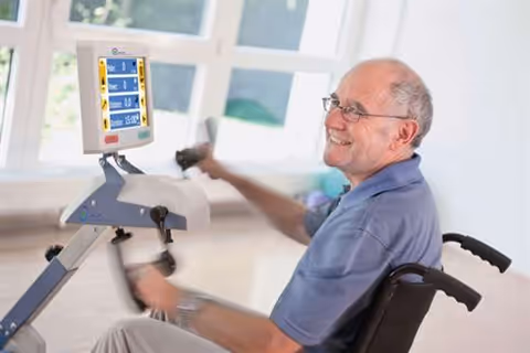 An elderly man in a wheelchair using an arm exercise machine with a digital display in a bright room with large windows.