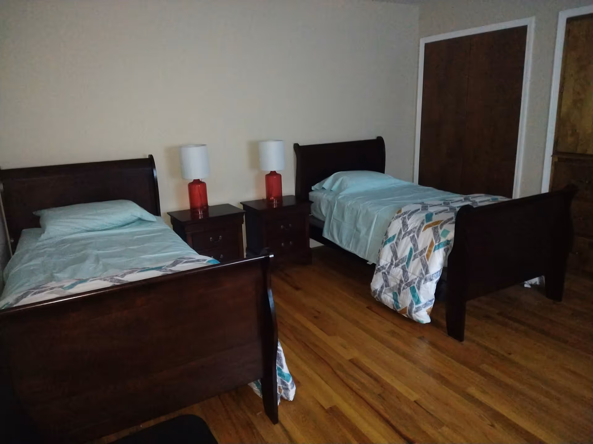 A bedroom with two single wooden beds, each made with light blue bedding and a patterned blanket at the foot. Between the beds are two dark wooden nightstands, each with a red lamp topped with a white lampshade. The room has hardwood floors and a closet with dark wooden sliding doors.