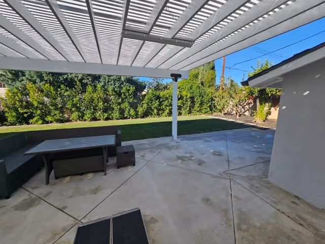 Outdoor patio area with a white pergola overhead, a black outdoor sectional sofa with a matching table and ottoman on a concrete floor. In the background, there is a green lawn bordered by tall bushes and trees under a clear blue sky.