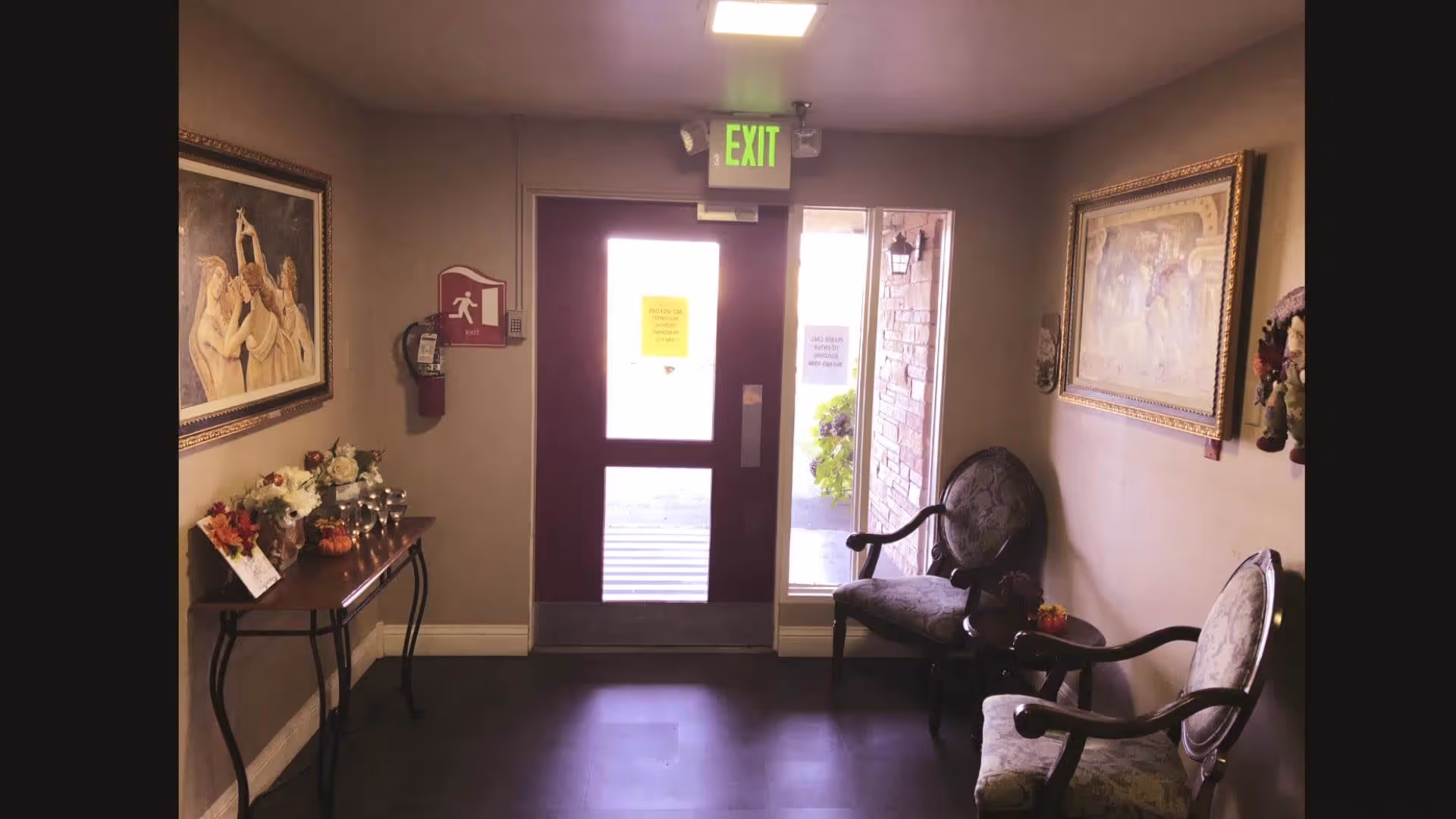 Small waiting area with two upholstered chairs and a small round table between them, a wooden console table with flower arrangements and decorative items, two framed paintings on the walls, and a glass exit door with an illuminated green exit sign above it.