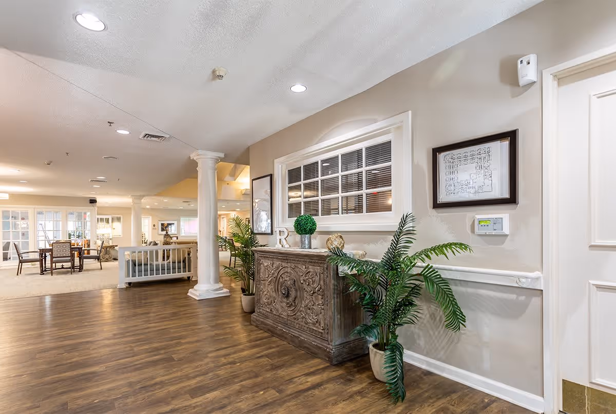 Interior view of a senior living facility hallway with wooden flooring, white columns, and beige walls. There is a decorative wooden cabinet with plants on either side, a framed floor plan on the wall, and a window with blinds above the cabinet. In the background, there is a seating area with chairs and tables.