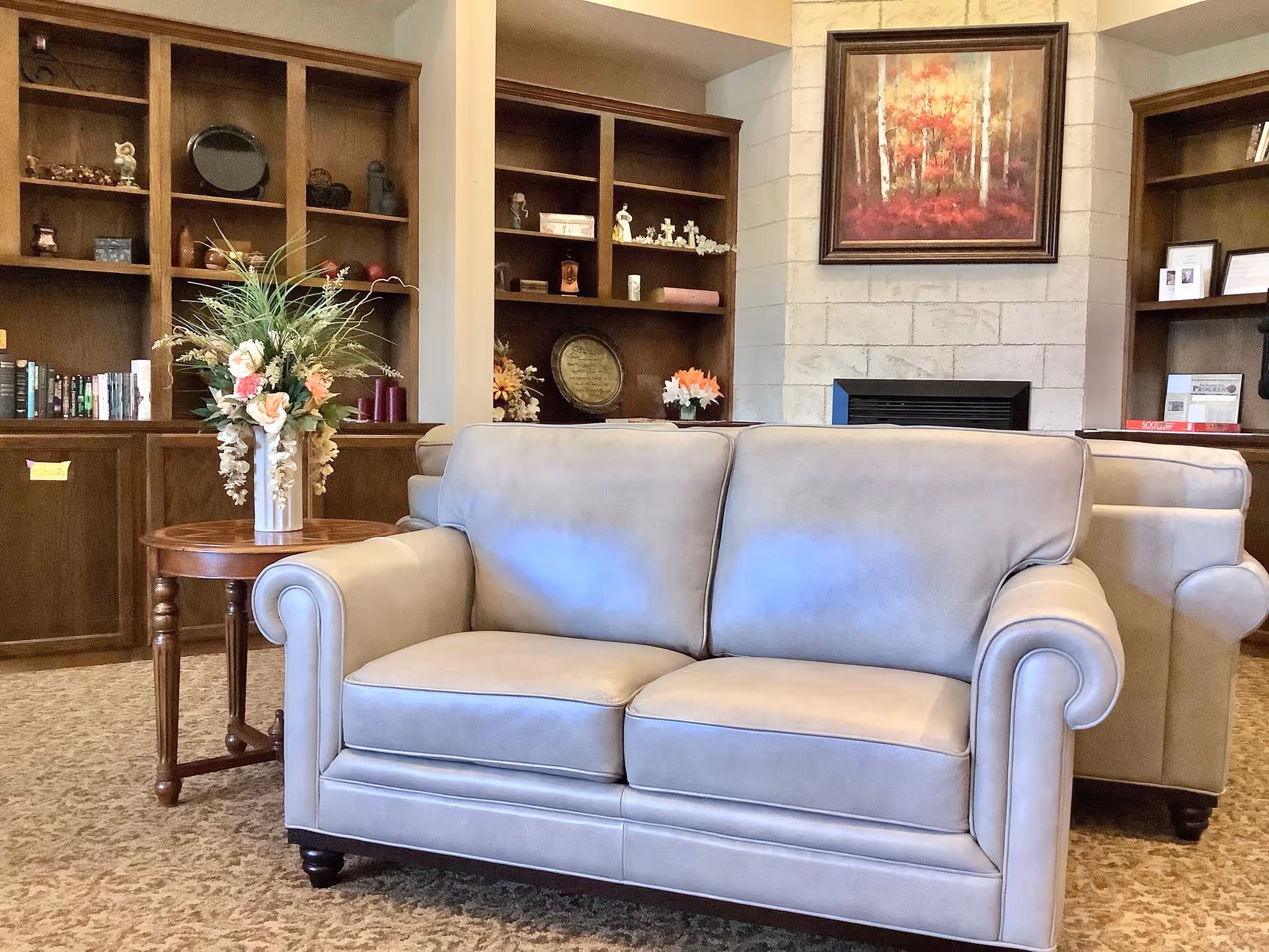A cozy living room area featuring two light gray leather sofas facing each other, a round wooden side table with a floral arrangement, built-in wooden bookshelves filled with decorative items and books, a stone fireplace, and a framed painting of a forest with autumn colors above the fireplace.
