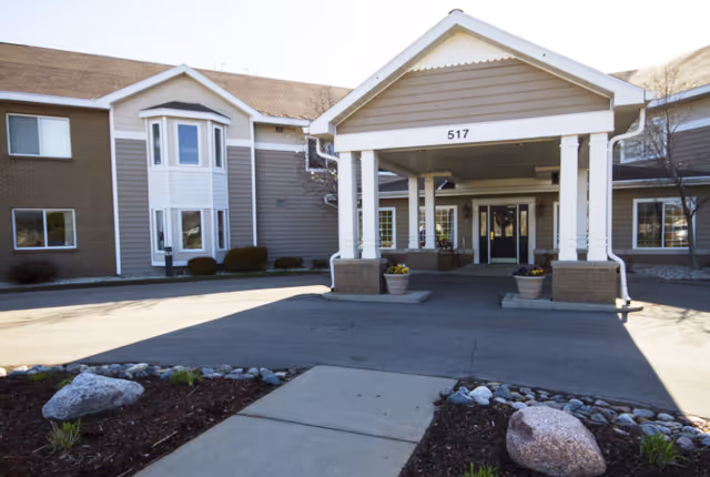 Front exterior view of a two-story senior living facility building with a covered entrance labeled 517, surrounded by landscaping with rocks and small plants.