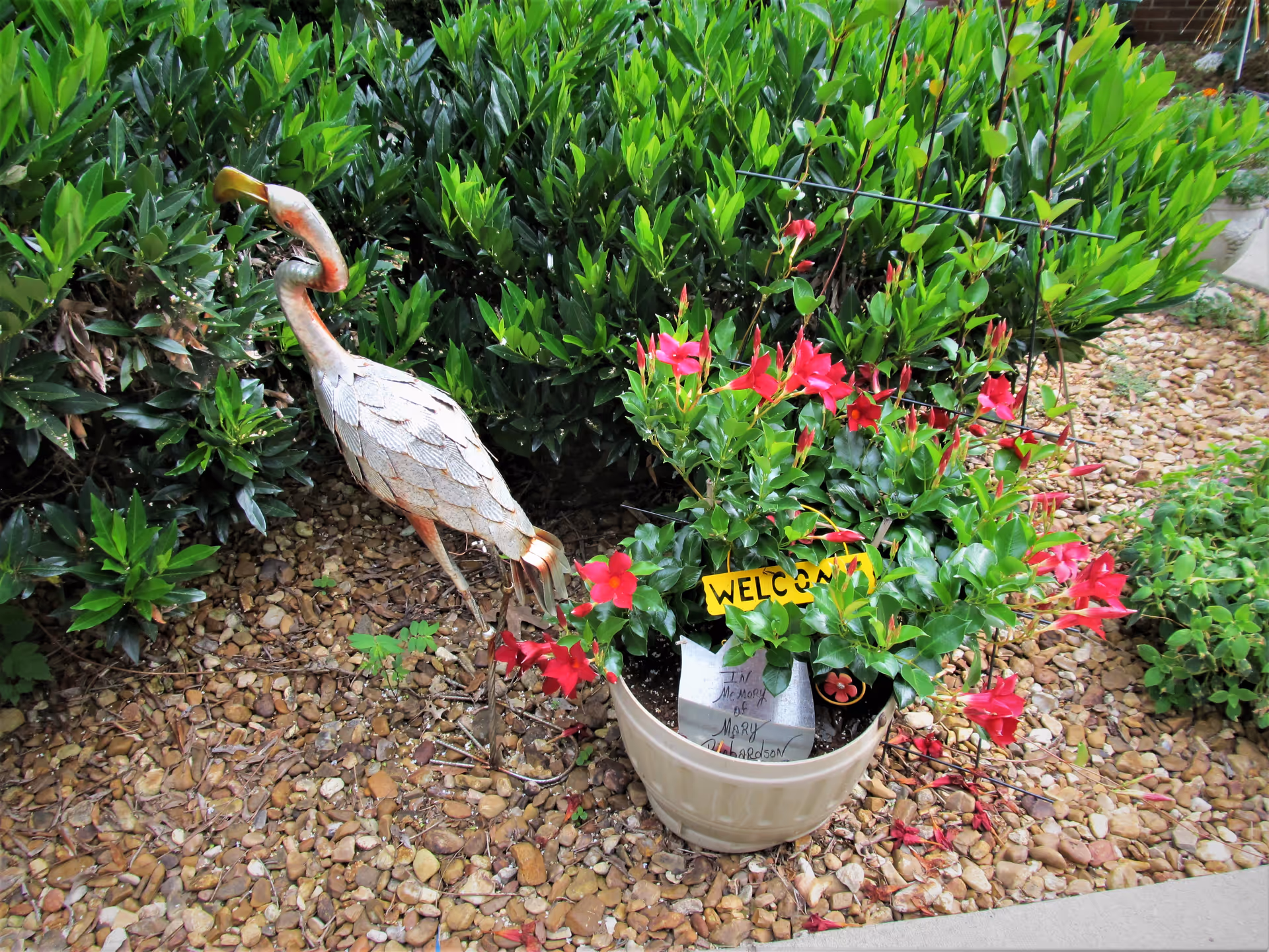 A garden area with a decorative metal bird sculpture standing on a bed of small rocks next to a potted plant with red flowers and a yellow 'WELCOME' sign. Green bushes are in the background.