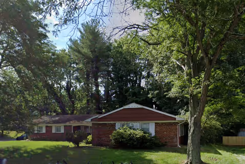 Single-story brick building with a sloped roof surrounded by green trees and grass under a clear sky.