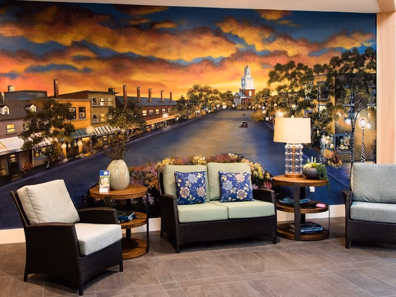 Comfortable seating area with wicker chairs and a loveseat placed in front of a large painted mural of a town at sunset.