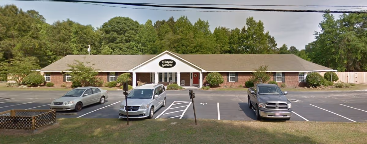 Front exterior view of a single-story brick building with a sign that reads 'Wisteria Gardens' above the entrance. There are several cars parked in the parking lot in front of the building, and trees and greenery surround the area.
