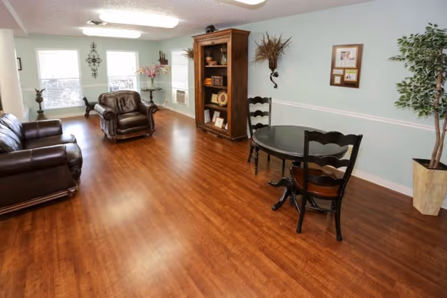 A spacious living room with wooden flooring, light blue walls, and white trim. The room features two dark brown leather sofas, a wooden bookshelf with decorative items, a small round black table with two chairs, a potted plant, and wall decorations including a framed picture and a wall-mounted floral arrangement. Large windows allow natural light to enter the room.
