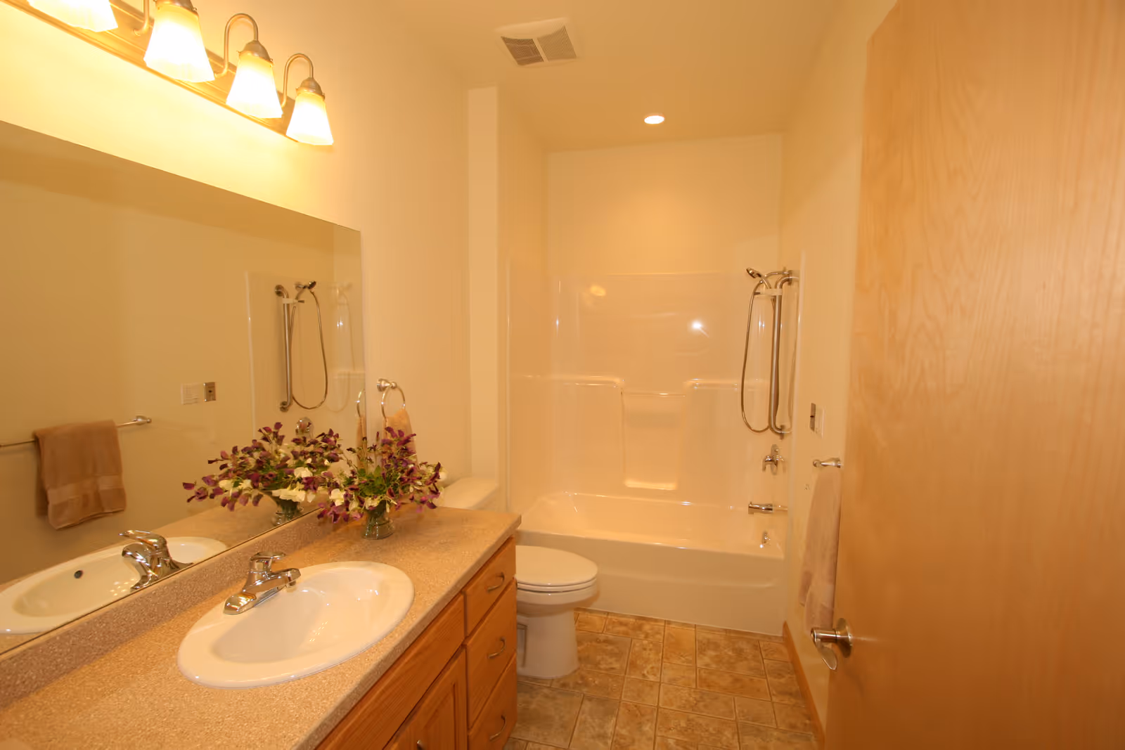 A clean bathroom with a single sink vanity topped with a beige countertop and a vase of purple and white flowers. Above the sink is a large mirror with three light fixtures. The bathroom has a white bathtub with a showerhead and grab bars, a toilet, beige tiled floor, and beige walls. A towel hangs on a rack next to the bathtub and another towel hangs on a rack near the sink.