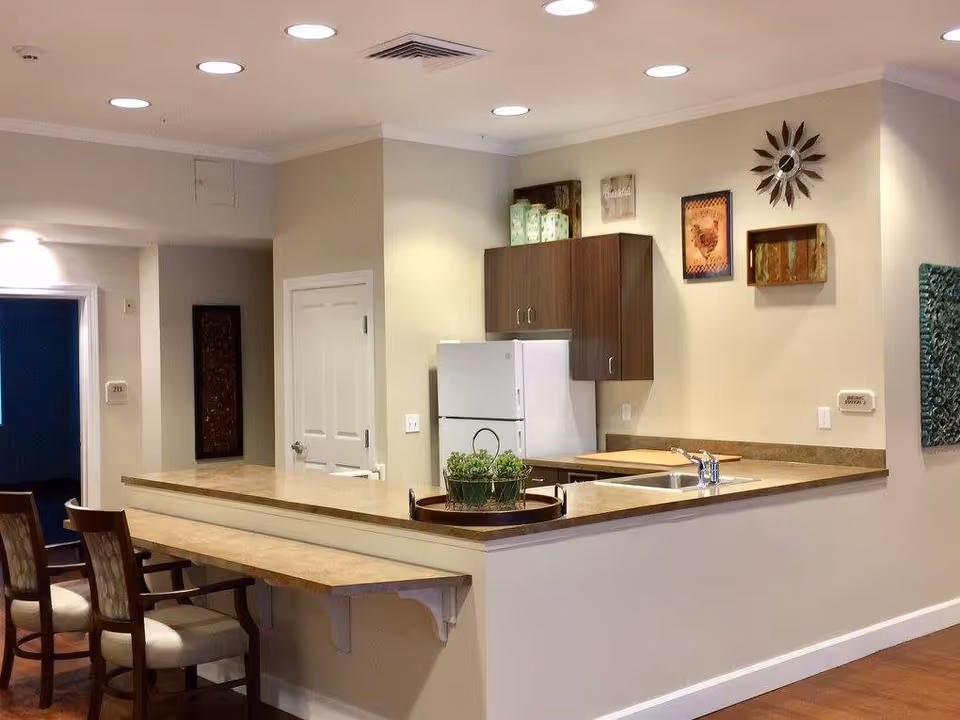 Interior view of a kitchen area in a senior living facility featuring a countertop with two chairs, a white refrigerator, wooden cabinets, a sink, and decorative wall art including a sunburst clock and framed pictures.