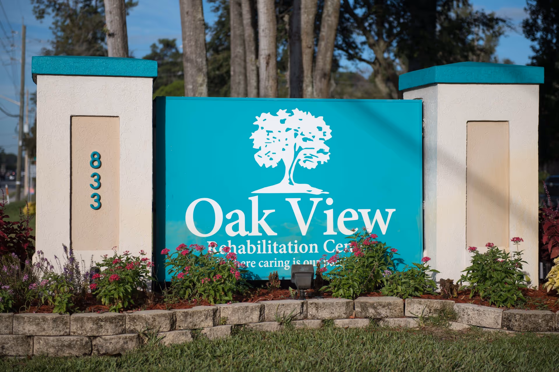 Outdoor sign for Oak View Rehabilitation Center with a white tree logo on a teal background, flanked by two beige pillars with teal tops and the number 833 on the left pillar, surrounded by flowers and greenery.
