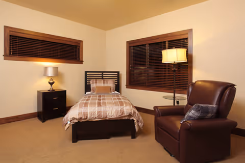 A cozy bedroom with a single bed featuring a checkered bedspread and a pillow, a dark wooden nightstand with a lamp, two windows with wooden blinds, a floor lamp, and a brown leather armchair with a plaid cushion.