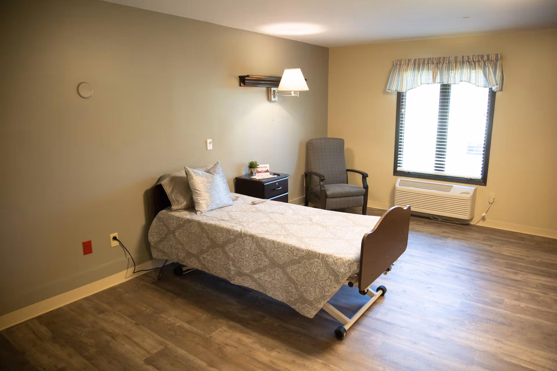 A simple, clean bedroom in a senior living facility with a single bed covered in a patterned bedspread, a nightstand with a small plant and a card, a wall-mounted lamp above the bed, a comfortable armchair near a window with blinds and a valance, and a wall-mounted air conditioning unit below the window. The room has wood flooring and beige walls.