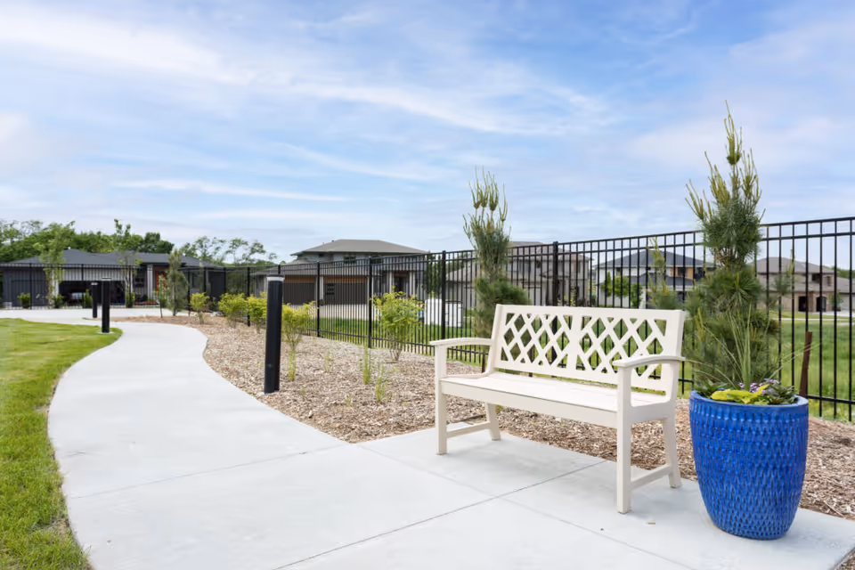 A curved concrete pathway in an outdoor garden area with a white bench and a large blue planter containing a small tree and flowers. The area is bordered by a black metal fence with houses and greenery visible in the background under a partly cloudy sky.