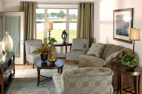 Sunlit living room with a sofa, patterned armchairs, coffee table and a large window overlooking a grassy area.