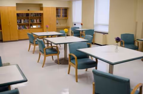 Interior dining area with multiple tables and teal chairs in a senior facility.