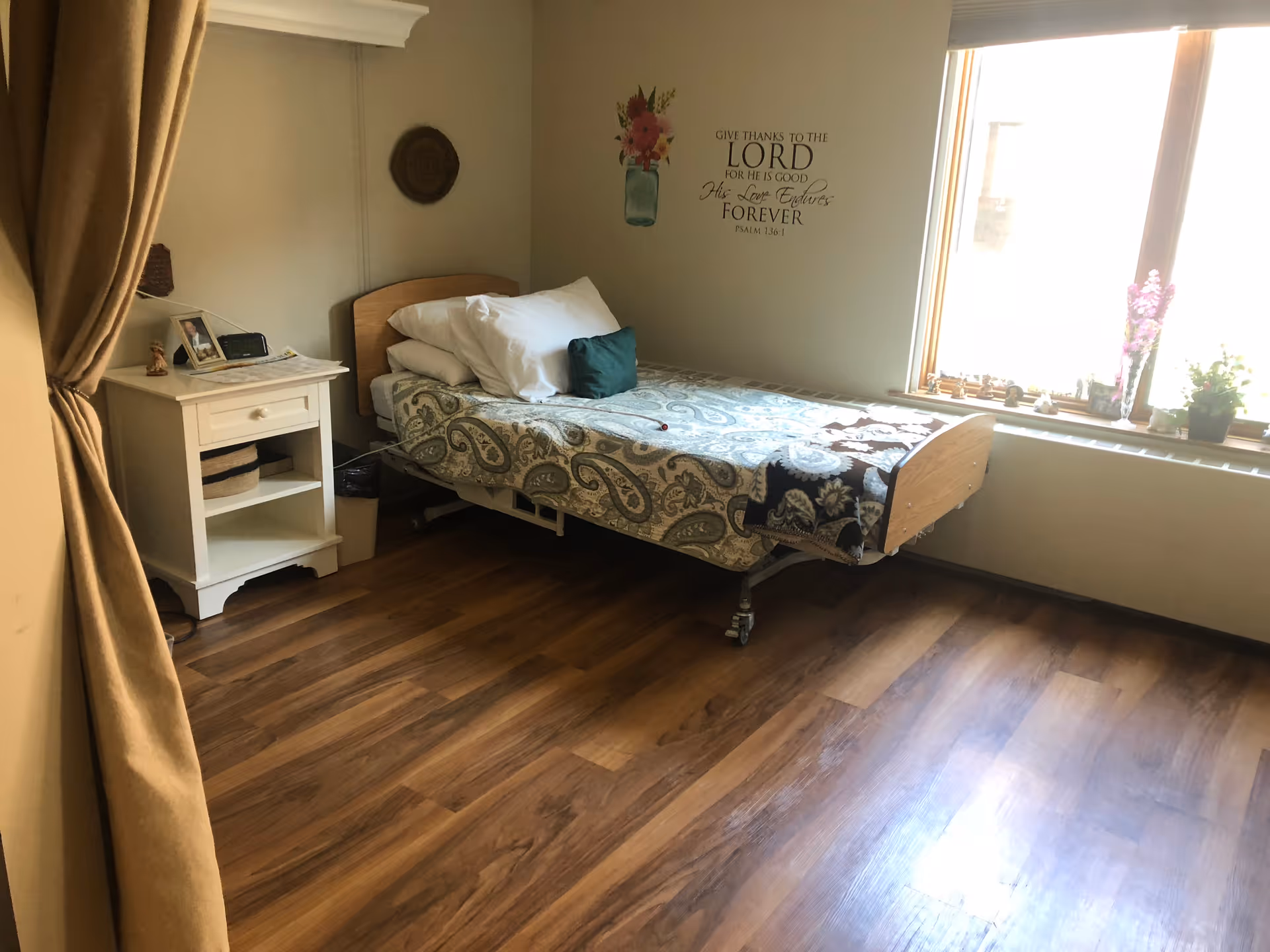 A single bed with patterned bedding and pillows is positioned against a wall in a room with wooden flooring. Next to the bed is a white nightstand with a drawer and open shelves, holding a photo frame, a clock, and some papers. A window with a windowsill decorated with small plants and figurines lets in natural light. On the wall above the bed is a decorative wall decal with a flower in a jar and a religious quote from Psalm 136:1. Beige curtains are partially visible on the left side of the image.