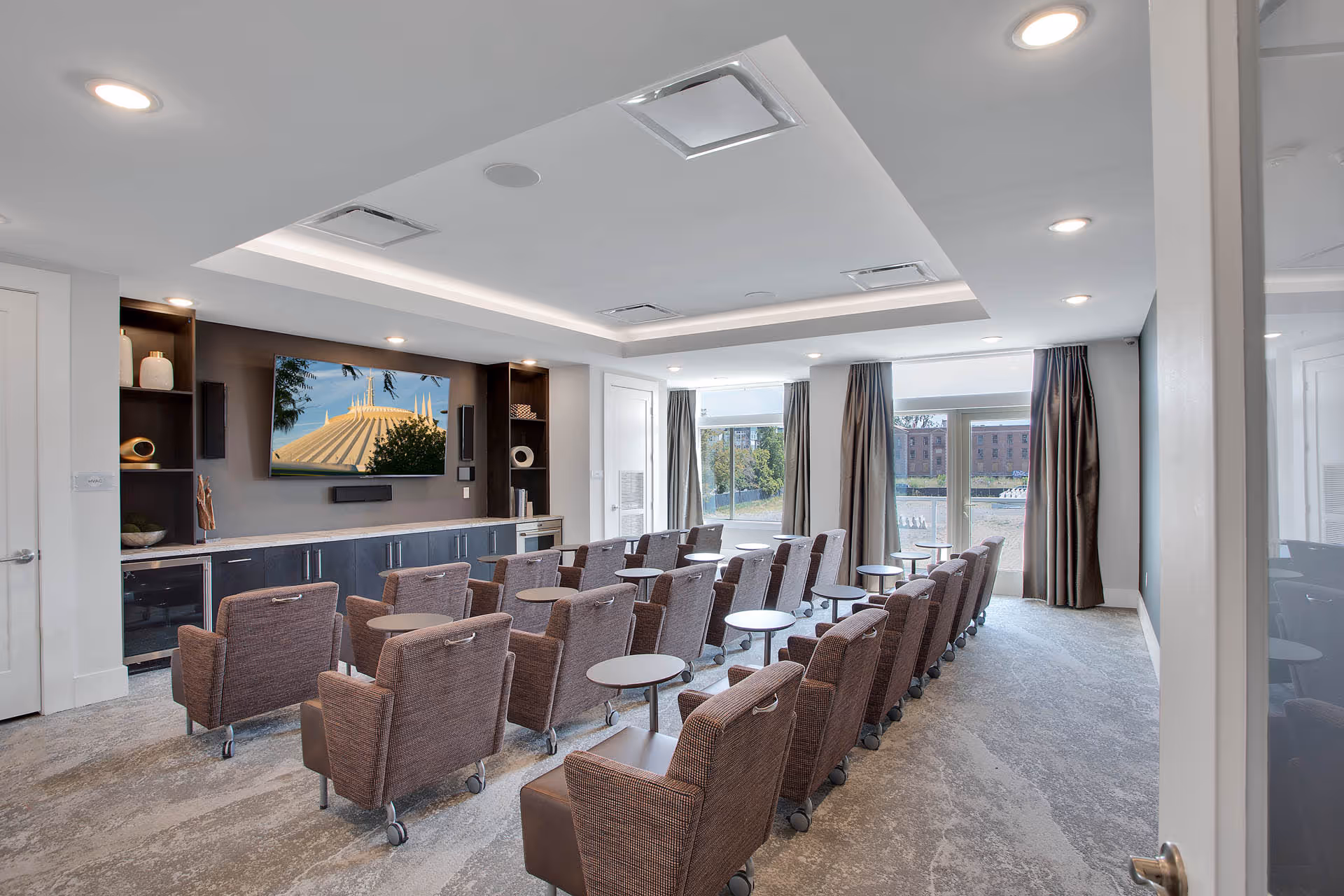 A modern media room with multiple rows of brown upholstered chairs on wheels, each paired with a small round side table. The room has a large flat-screen TV mounted on a wall with built-in shelves and cabinets below. Large windows with curtains allow natural light to enter, and the ceiling features recessed lighting and a tray design.
