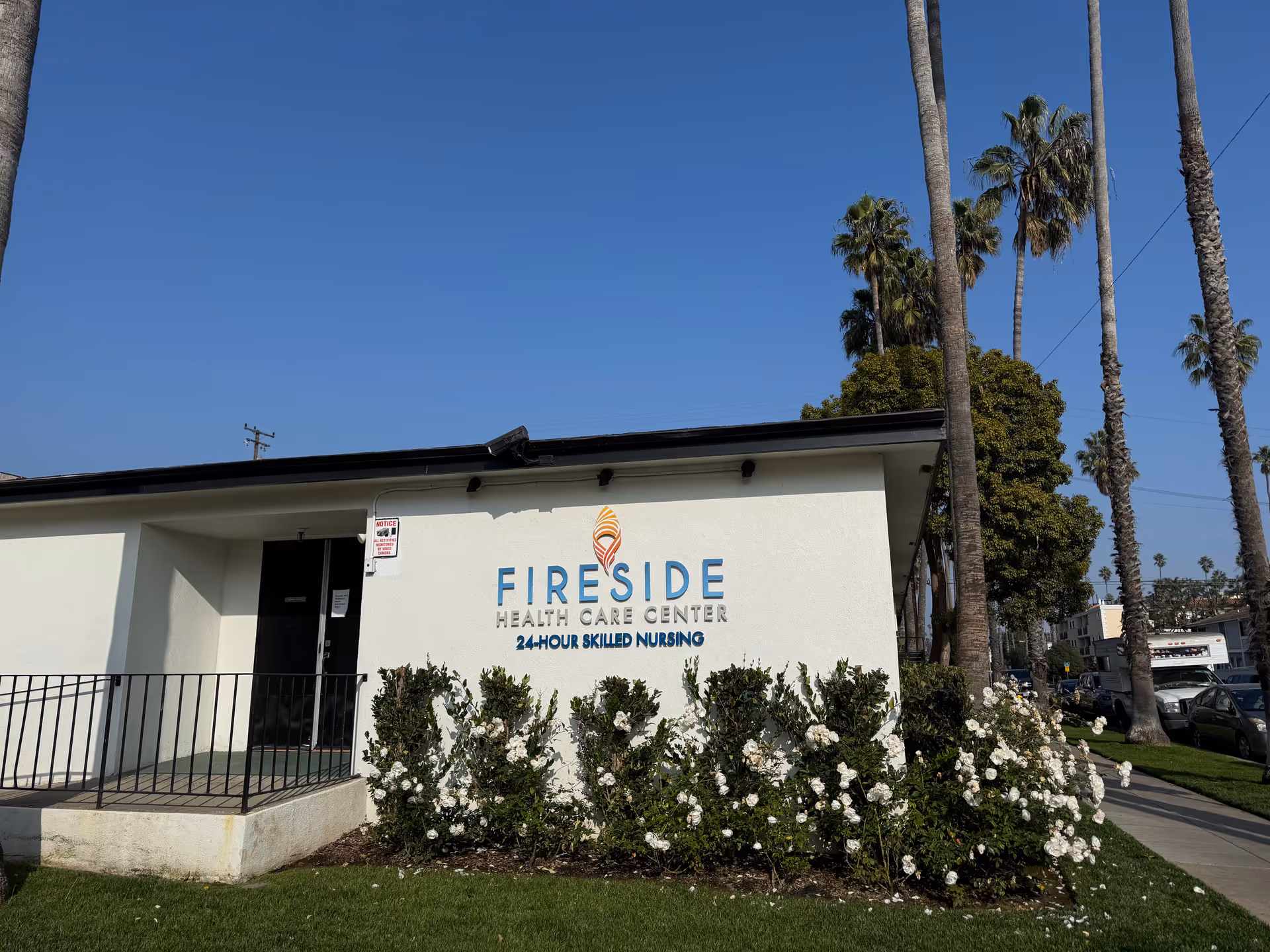 Exterior view of Fireside Health Care Center building with a white facade, a black door, and a sign that reads 'Fireside Health Care Center 24-Hour Skilled Nursing'. There are palm trees and white flowering bushes in front of the building under a clear blue sky.
