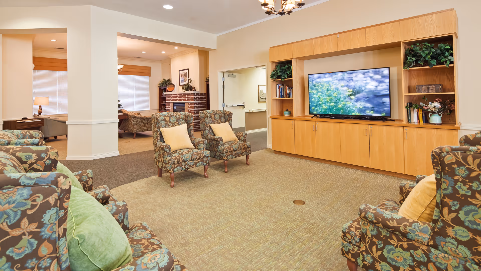 A spacious living room area in a senior living facility with multiple floral-patterned armchairs arranged around a large wooden entertainment center with a flat-screen TV. The room has beige walls, carpeted floors, and recessed lighting. There is a fireplace and additional seating visible in an adjoining room.