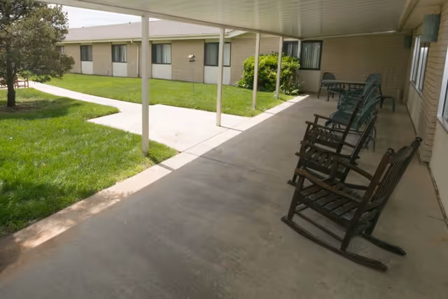 Covered outdoor walkway and courtyard with a row of wooden rocking chairs and patio tables beside a grassy lawn and single-story building.