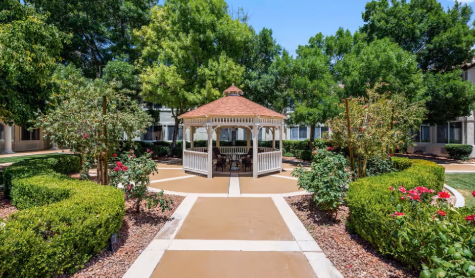 A landscaped outdoor garden area with a paved walkway leading to a white gazebo surrounded by green bushes, flowering plants, and trees. The background shows parts of a building with windows.