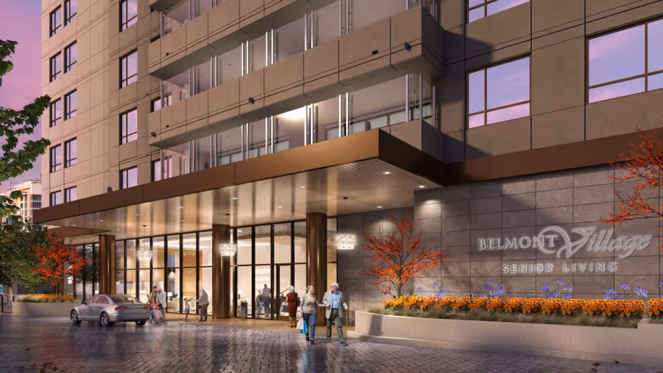 Exterior view of Belmont Village Senior Living La Jolla building entrance at dusk with people walking and a car parked nearby. The building has large glass windows and a modern design with landscaping including flowers and small trees.