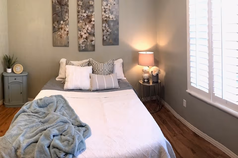 A cozy bedroom with a neatly made bed featuring multiple pillows and a light blue throw blanket. There is a small round side table with a lamp and a flower vase on the right side of the bed, and a small cabinet with a plant and a decorative plate on the left side. Three floral paintings hang on the wall above the bed, and a window with white shutters is on the right wall, allowing natural light to enter the room.