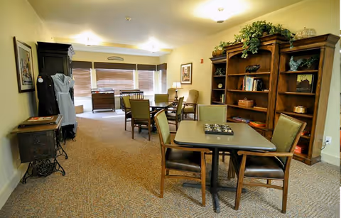 A cozy common area with a table and four chairs in the foreground, a large wooden bookshelf filled with books and decorative items on the right, and additional seating and furniture near large windows with blinds in the background. The room is warmly lit with ceiling lights and decorated with plants and framed pictures.