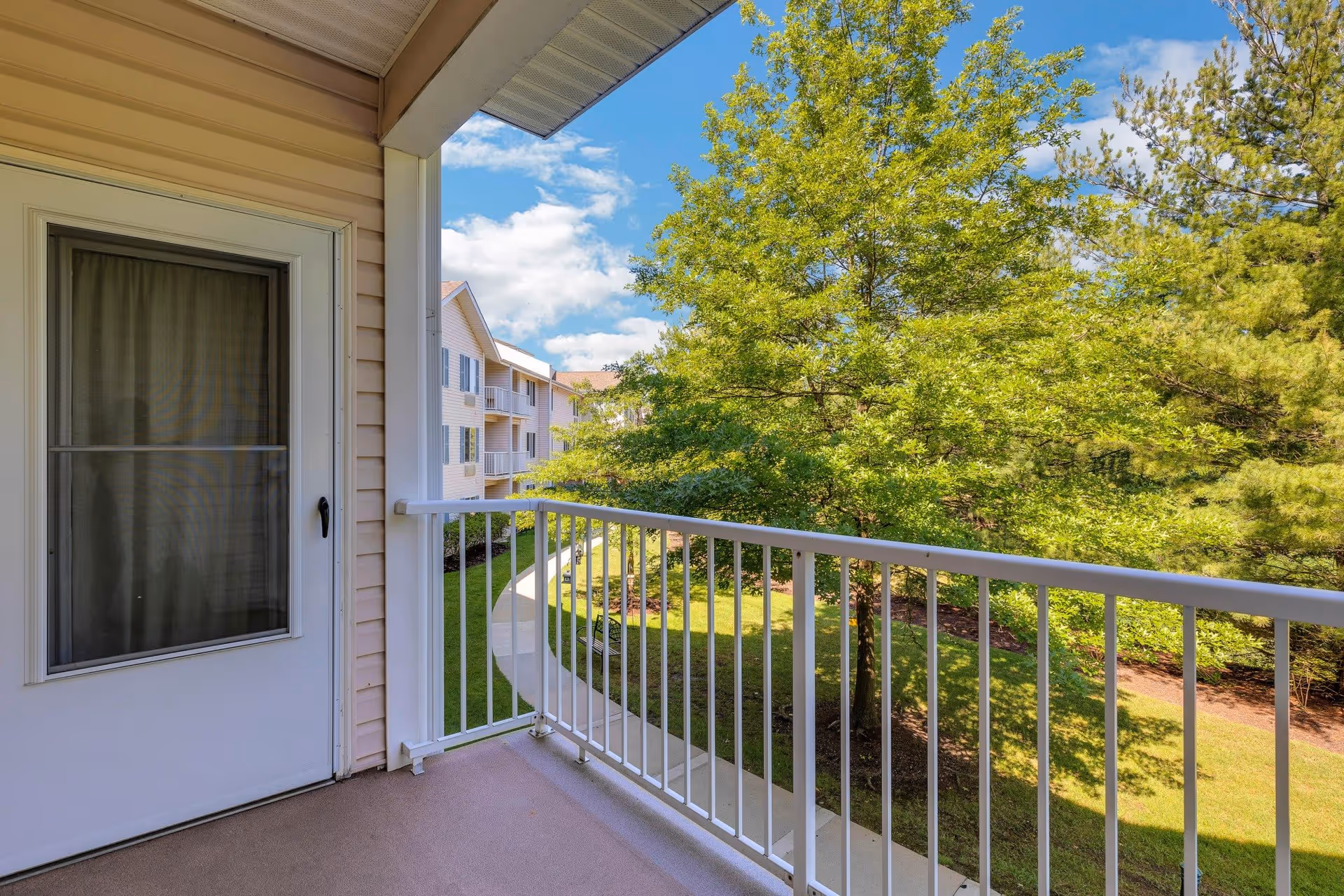 View from a balcony with white railing overlooking a green landscaped area with trees and a curved sidewalk. Part of a beige building with a white door and window is visible on the left side under a partly cloudy blue sky.