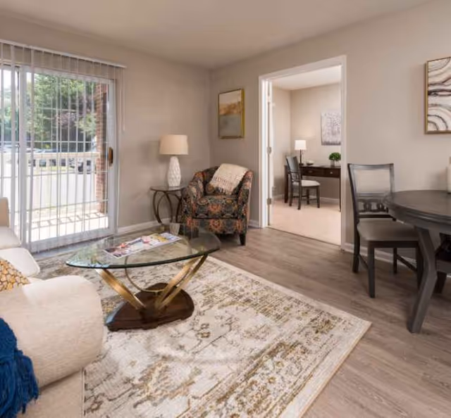 A cozy living room with a beige sofa, a patterned armchair, a glass coffee table with a wooden base, and a floor lamp on a small side table. The room has a large window with vertical blinds letting in natural light. Adjacent to the living room is a small office area with a desk, chair, and lamp. The flooring is wood with a decorative area rug.