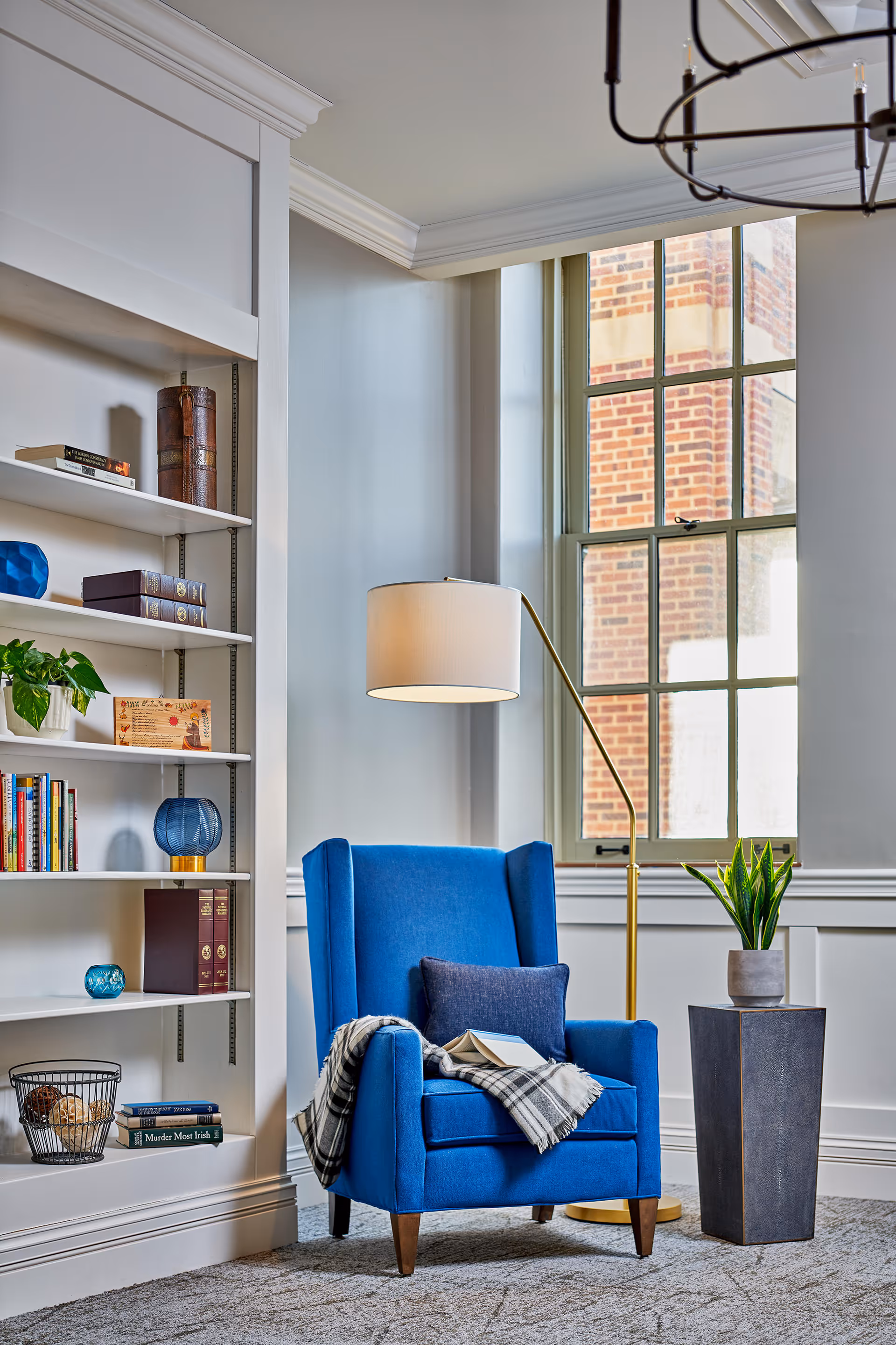 A cozy reading nook featuring a bright blue armchair with a dark blue pillow and a plaid throw blanket draped over one arm. An open book rests on the chair. Next to the chair is a tall gold floor lamp with a white lampshade and a tall planter with a green plant. Behind the chair is a large window with a view of a brick building. To the left, there is a white built-in bookshelf with various books, decorative items, and a small plant.