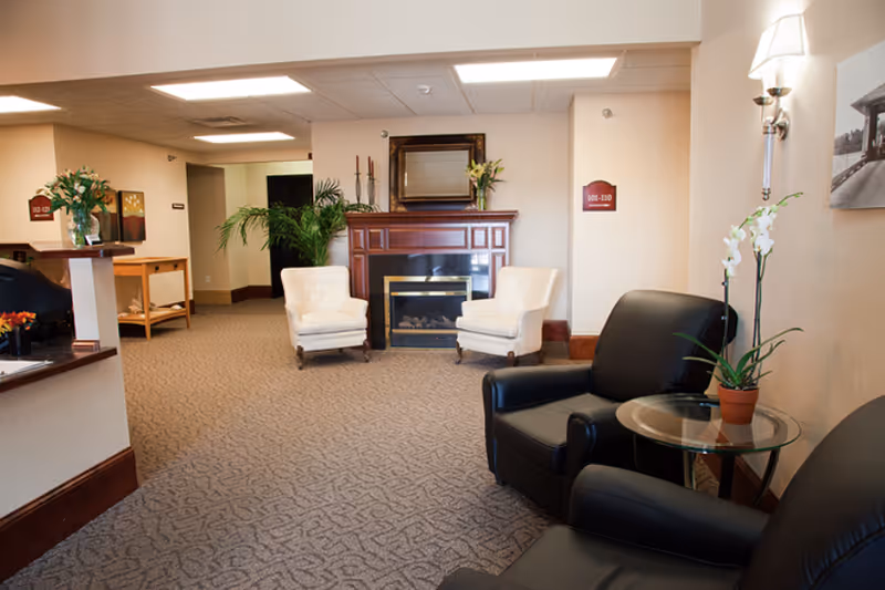 A comfortable senior living facility common area with armchairs, a fireplace, and potted plants.