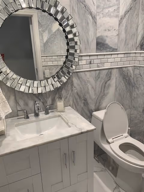 A modern bathroom with marble-patterned walls and a white vanity with a marble countertop. There is a rectangular sink with a chrome faucet, a soap dispenser, and a round decorative mirror with a geometric frame above the sink. To the right, there is a white toilet with the lid open.