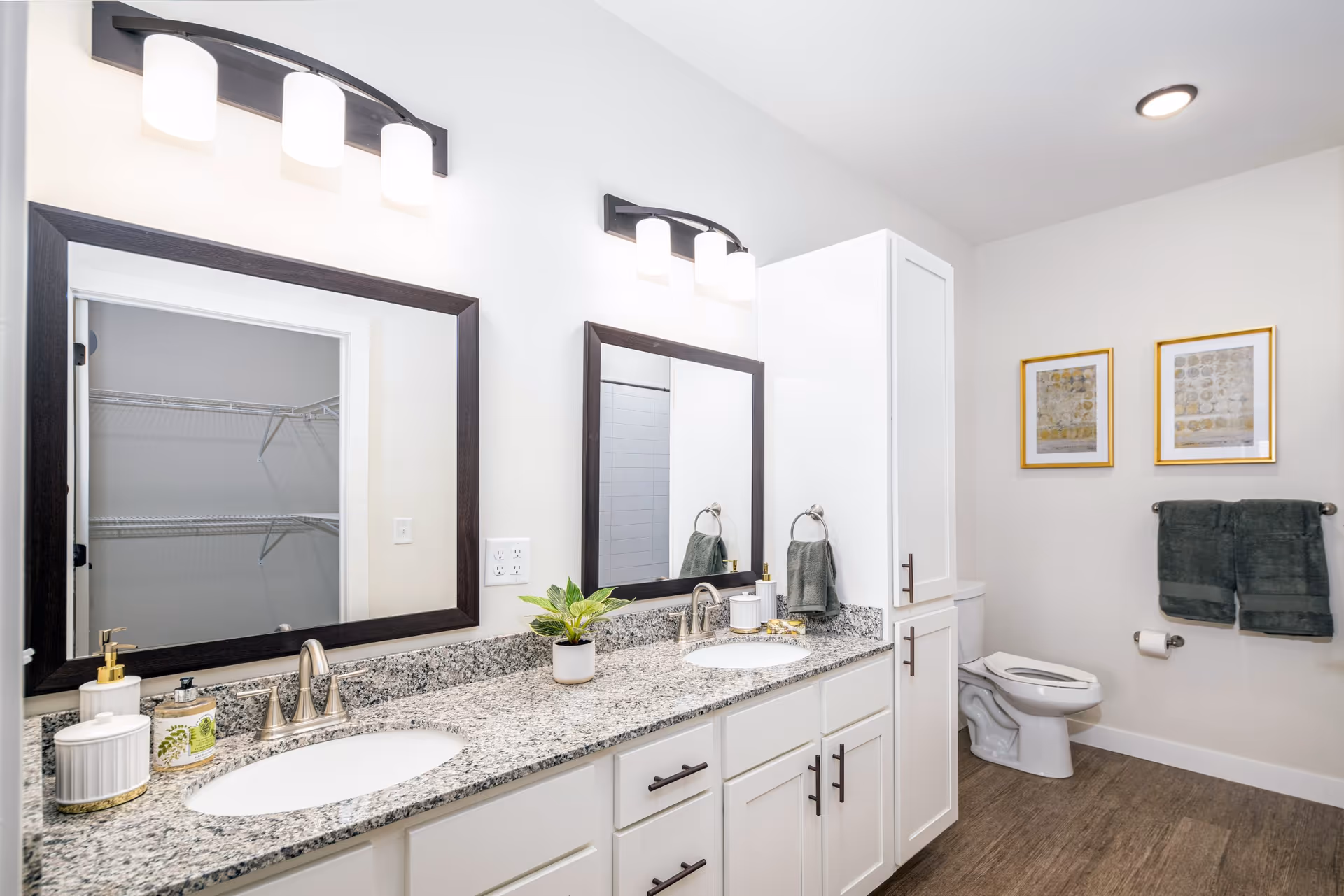 Bright and modern bathroom with a double sink vanity featuring granite countertops, two large mirrors with dark frames, and overhead lighting. There are white cabinets below the sinks and a tall white cabinet between the sinks and the toilet. The toilet is positioned near the wall with two framed artworks above it and two dark green towels hanging on a towel rack. The floor has a wood-like finish.