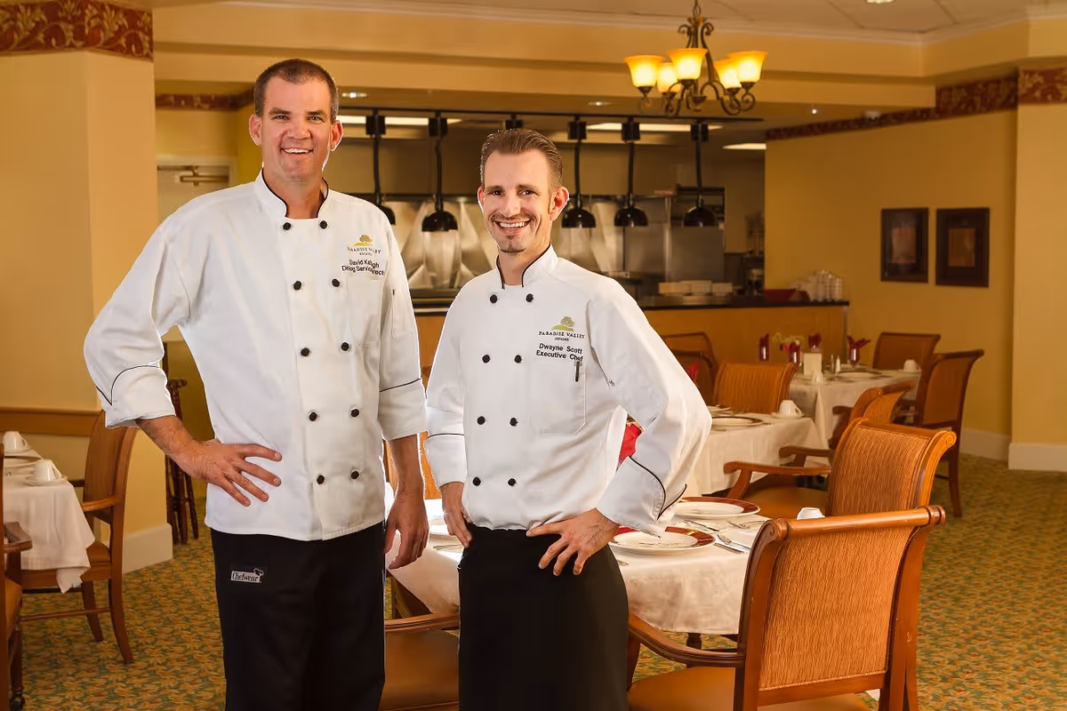 Two male chefs standing and smiling in a dining room with tables set for a meal. Both are wearing white chef jackets with the Paradise Valley Estates logo and black pants. The dining room has warm lighting, wooden chairs, and tables covered with white tablecloths.