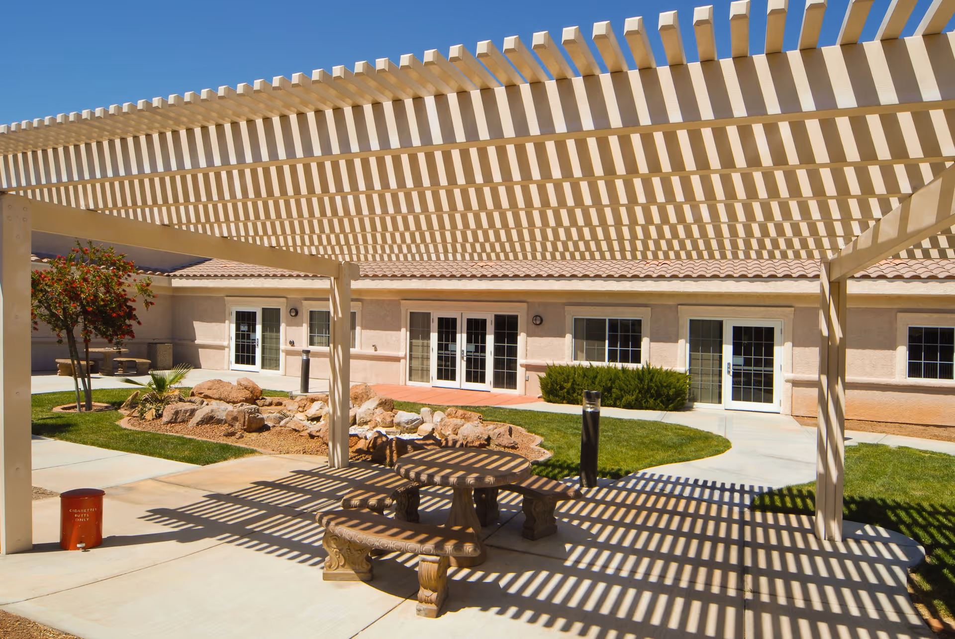 Outdoor seating area at Life Care Center of South Las Vegas with a pergola casting striped shadows over a round stone table and benches. The area is surrounded by a landscaped garden with rocks, grass, and small trees, and the building with multiple glass doors and windows is visible in the background under a clear blue sky.
