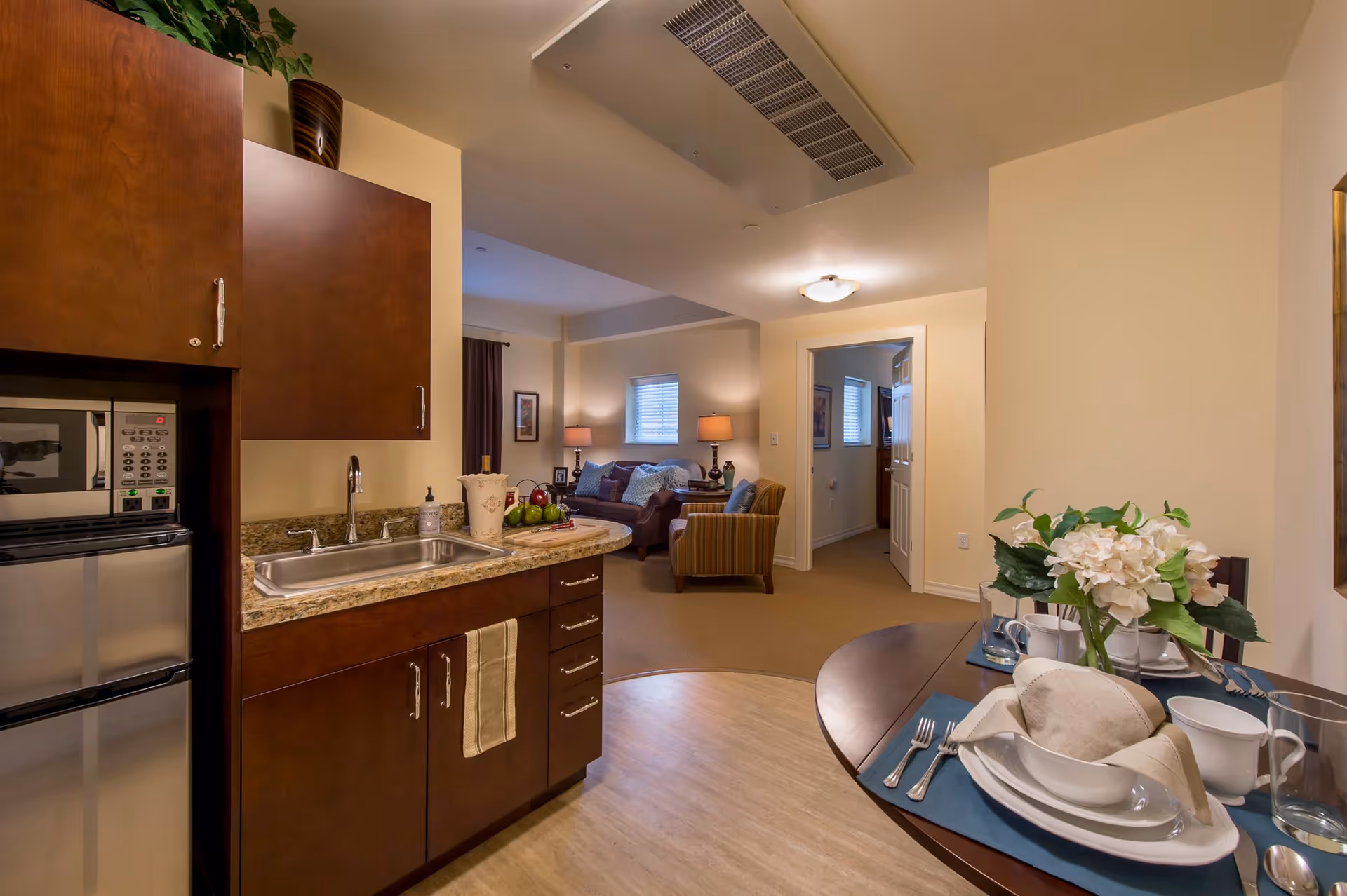 View of a small kitchen area with wooden cabinets, a sink, microwave, and mini refrigerator. Adjacent to the kitchen is a dining table set with plates, bowls, cups, and silverware, decorated with a floral centerpiece. In the background, there is a living room with a sofa, armchair, lamps, and framed pictures on the walls. A hallway leads to another room with a door partially open.
