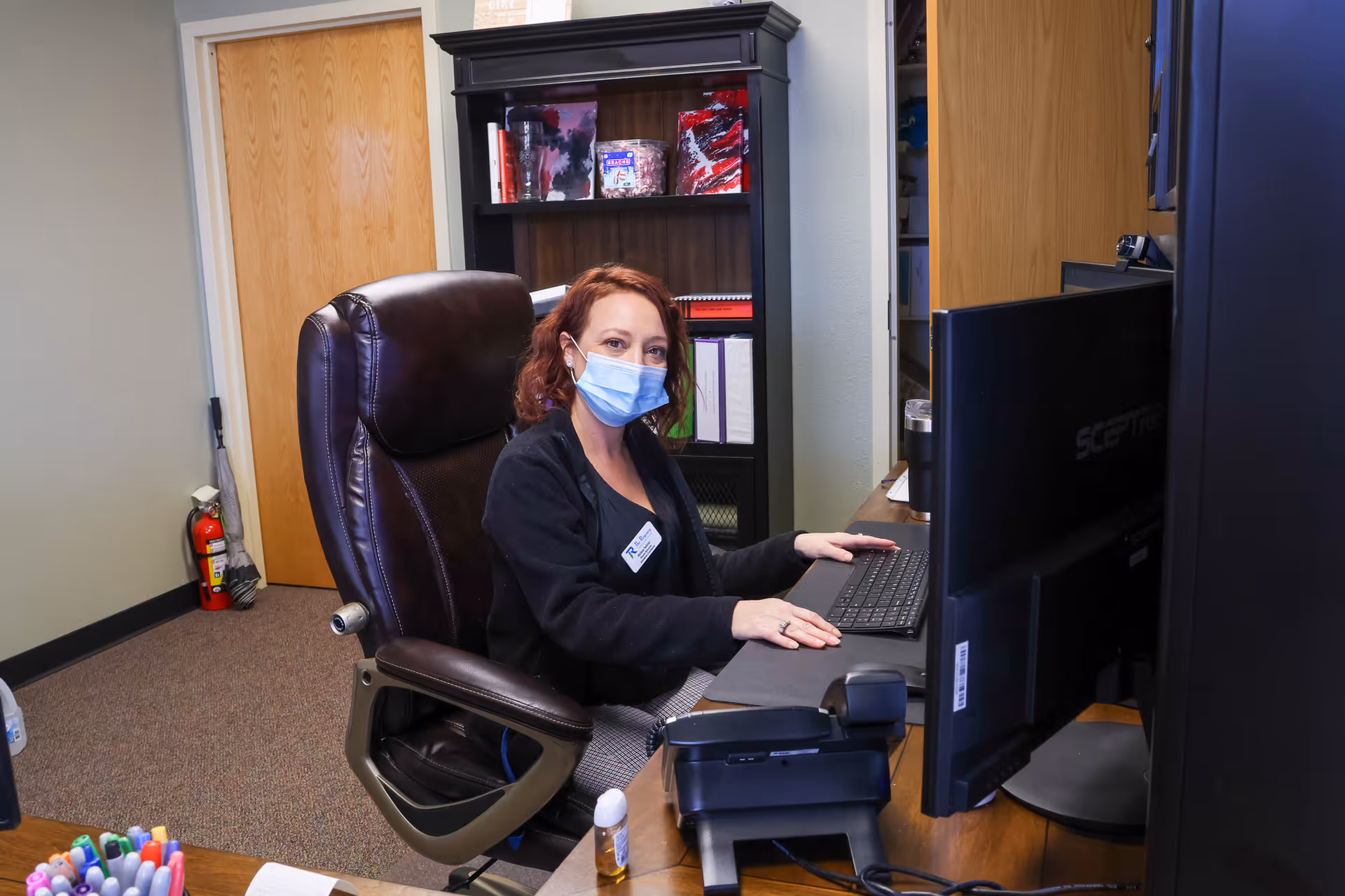 A woman wearing a face mask sits in a leather office chair at a desk with a computer keyboard and monitor. Behind her is a wooden bookshelf with various items including binders and decorative containers. The room has beige walls, a wooden door, and a fire extinguisher near the door.
