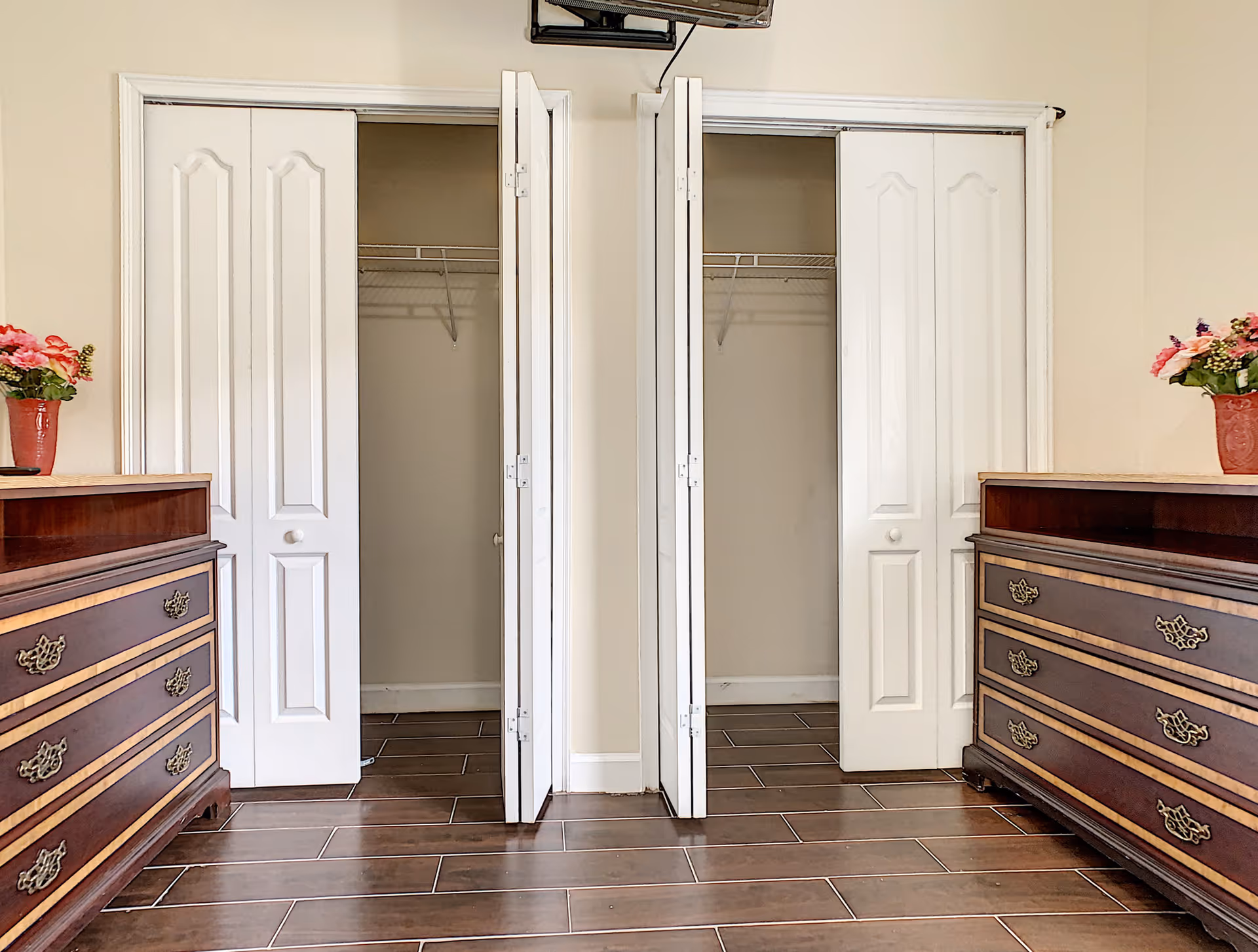 A bedroom interior showing two open closets flanked by matching wooden dressers with flower vases and wood-look tile flooring.